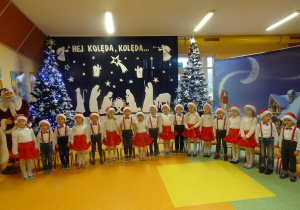 Grupa dzieci śpiewa kolędę, w tle dekoracja świąteczna, choinki.